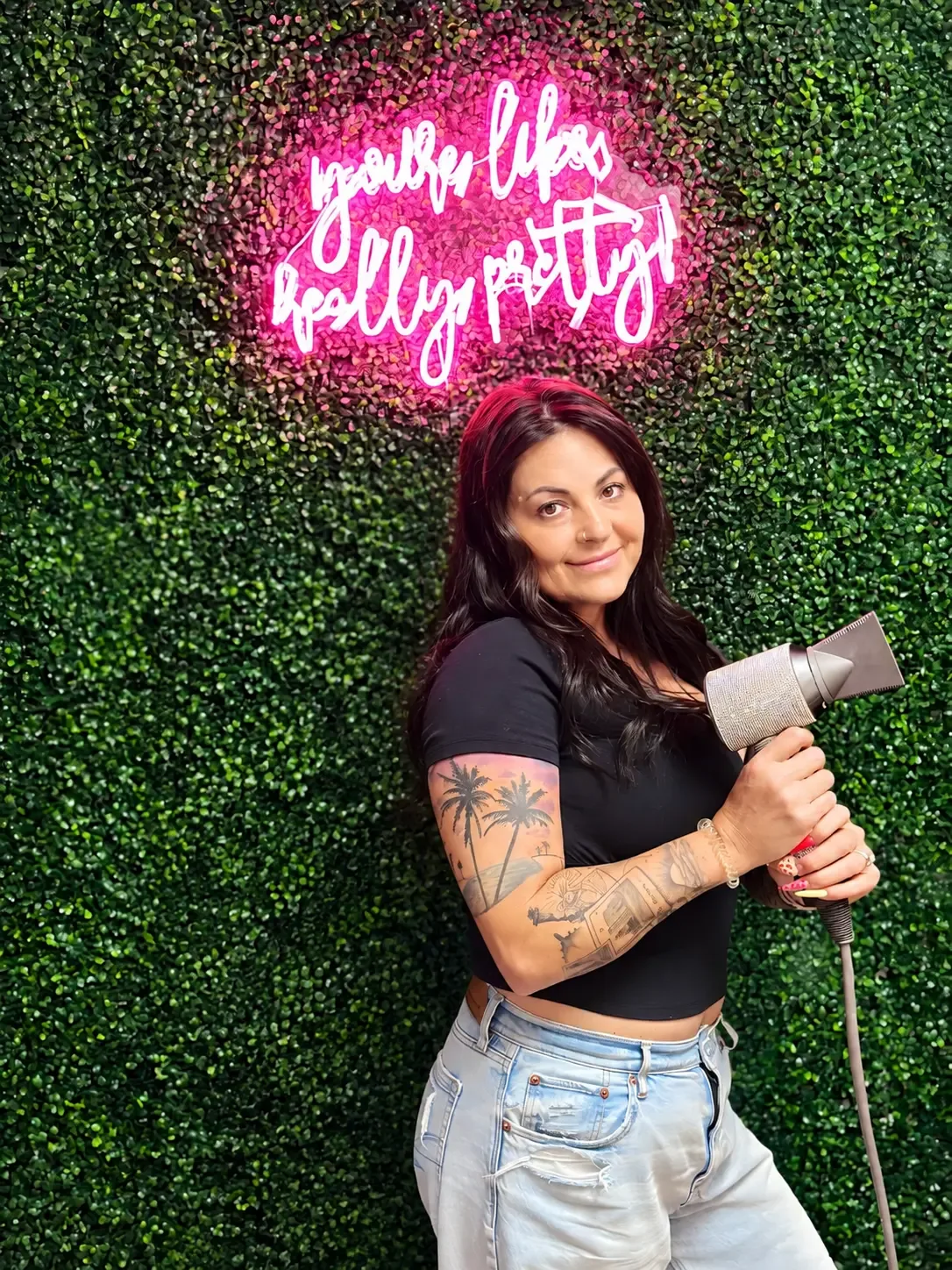 Jennifer, stylist at JM Salon in Lake Park, holding a hair dryer by the salon greenery wall